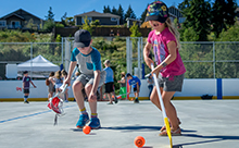 Two children play ball hockey and lacrosse in the foreground on an outdoor rink surface in summer.