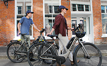 Two people walk alongside their CRD electric bicycles in a sidewalk.
