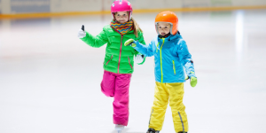 Two children in snow pants, jackets and helmets skate on an indoor ice rink.