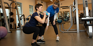 A SEAPARC weight room employee stands next to a patron, instructing her in a weighted squat with a kettle bell.