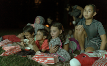 A crowd of kids sit and lie down on the grass, watching a movie playing in an outdoor theatre.