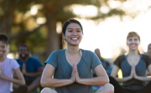 A woman smiling while holding child's pose during an outdoor yoga class, with fellow students in the background.